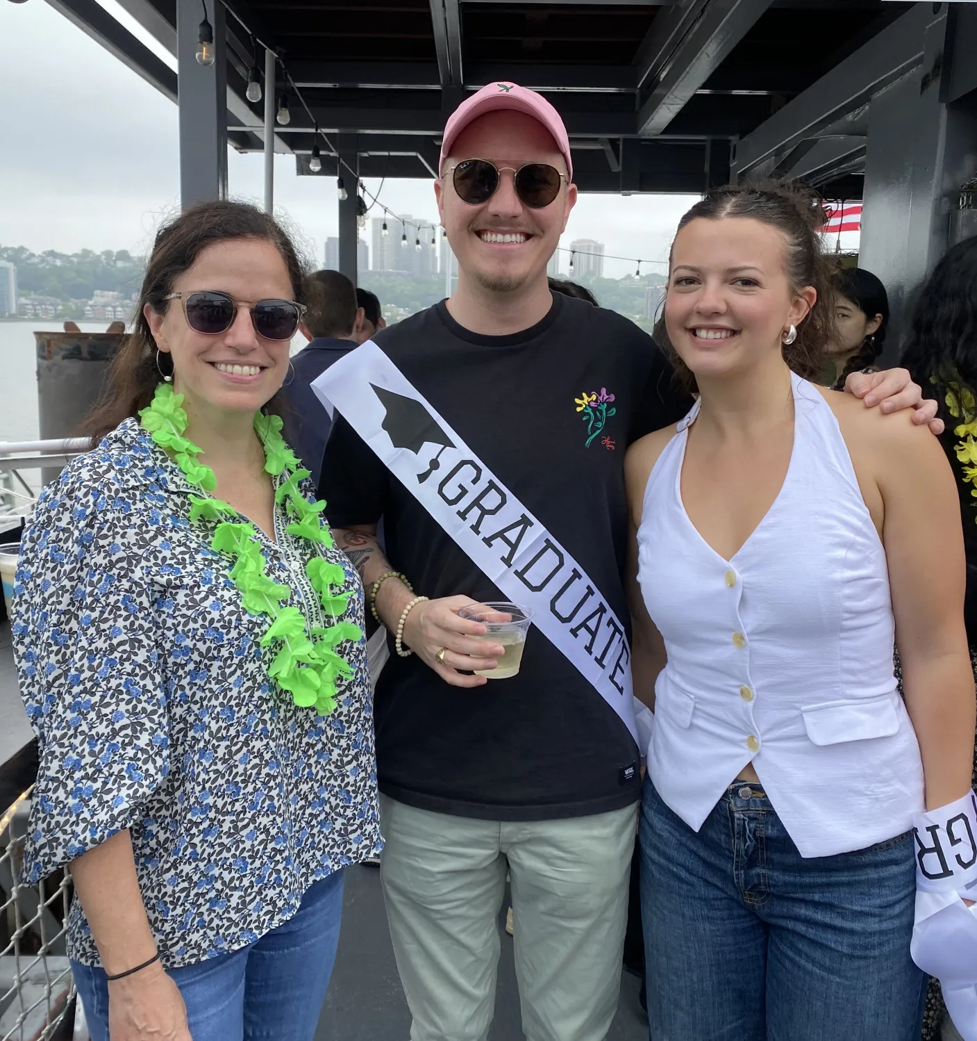 Alec Batts and Maggie Elizabeth Nedbalek stand smiling with Professor Elisa Konofagou aboard a boat during the Columbia BME 2025 Department Celebration. Alec wears a black shirt and pink cap with a ‘Graduate’ sash, Maggie is in a white sleeveless top and jeans, and Elisa wears sunglasses and a patterned shirt with a green lei. The Hudson River and cityscape are visible in the background under overcast skies.